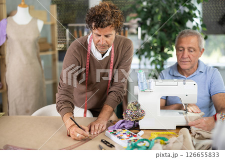 Elderly man sews on machine male assistant drawing pattern Elderly man sews on machine male assistant drawing pattern 126655833