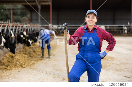 Hardworking young woman farmer in overalls posing with rake and smiling at camera while working on cow farm 126656024