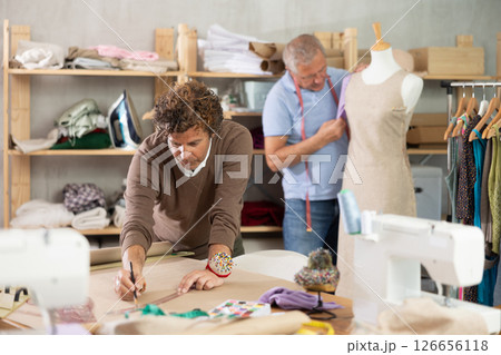 Adult man drawing pattern on paper male assistant sews on mannequin Adult man drawing pattern on paper male assistant sews on mannequin 126656118