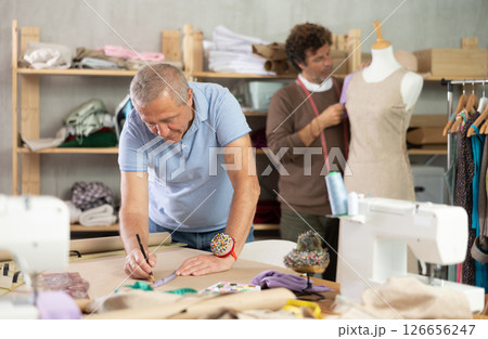 Senior man tailor draw dress pattern of product on paper, other work with mannequin. Senior man tailor draw dress pattern of product on paper, other work with mannequin. 126656247