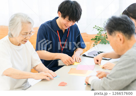 グループホーム,老人ホームで折り紙をする高齢者と介護士の男女スタッフ 126657600