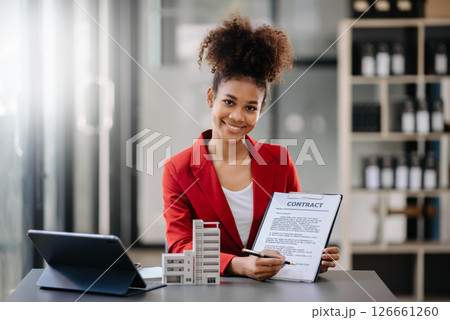 Young real estate agent worker working with laptop and tablet at table in office and small house beside it. Young real estate agent worker working with laptop and tablet at table in office and small house beside it. 126661260