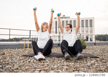 Senior man and woman sitting outdoors in rooftop area lifting dumbbells wearing casual fitness outfits promoting health and fitness. Visible joy and energy in performing physical exercise together 126661780