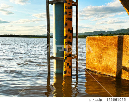 Water level at the dam, A water gauge stick in the middle of a river, showcasing accurate water level measurement with a scenic backdrop of mountains and cloudy sky Water level at the dam, A water gauge stick in the middle of a river, showcasing accurate water level measurement with a scenic backdrop of mountains and cloudy sky 126661936