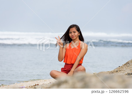 Happy young woman showing peace sign while sitting on beach 126662596