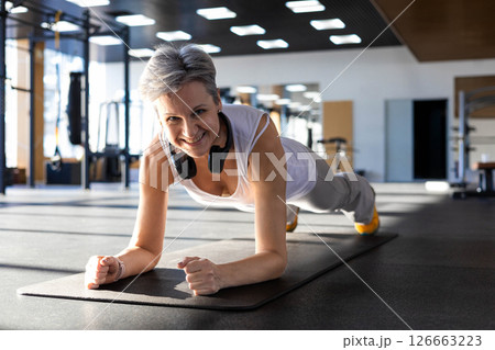 Smiling fitness An elderly gray-haired senior stand in plank position doing push ups training at gym 126663223