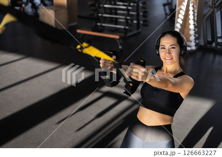 Young beautiful Asian woman doing fitness exercises on the TRX machine in the gym. Young beautiful Asian woman doing fitness exercises on the TRX machine in the gym. 126663227