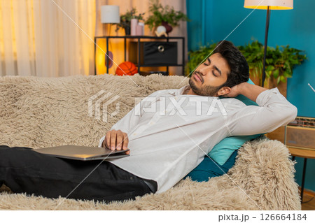 Indian young man falling asleep on sofa with laptop on lap, exhausted from work, tired and drained Indian young man falling asleep on sofa with laptop on lap, exhausted from work, tired and drained 126664184