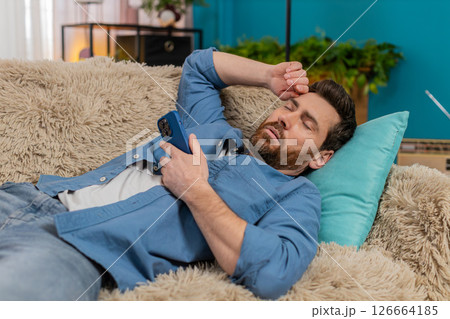 Young man unexpectedly falling asleep on sofa while holding smartphone in hand, tired and exhausted Young man unexpectedly falling asleep on sofa while holding smartphone in hand, tired and exhausted 126664185