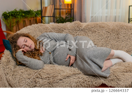 Pregnant young woman falling asleep on home sofa gently holding belly relaxed calm peaceful rest 126664337