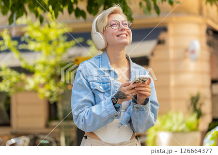 Overjoyed blonde woman in wireless headphones choosing, listening music holding smartphone on street 126664377