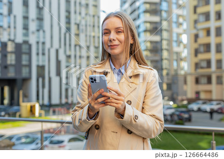 Happy mature businesswoman using smartphone and looking at camera while standing on downtown street 126664486