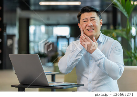 Chinese businessman in office lobby working on laptop and feeling toothache in modern office lobby 126665271