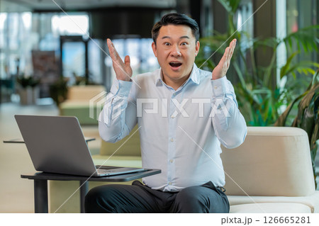 Chinese young businessman in shirt looking at camera shocked by sudden victory in office lobby 126665281