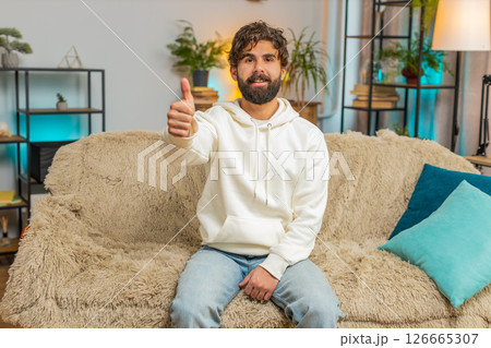 Happy excited Indian young man sitting on sofa couch looking at camera and showing thumbs up sign Happy excited Indian young man sitting on sofa couch looking at camera and showing thumbs up sign 126665307
