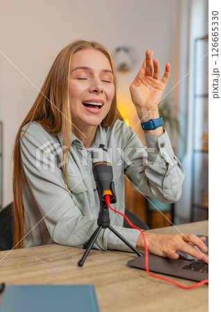 Mature positive Caucasian woman blogger singer using laptop while singing into microphone at table 126665330