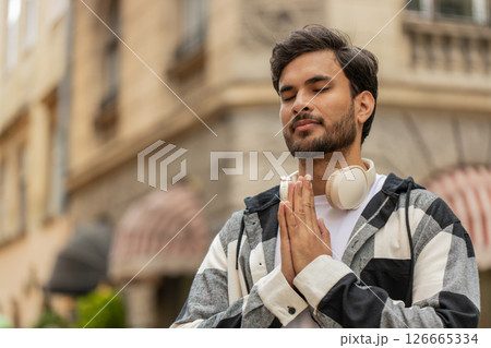 Religious Indian man praying with closed eyes to God asking for blessing while standing on street Religious Indian man praying with closed eyes to God asking for blessing while standing on street 126665334