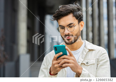 Indian man using smartphone typing text messages, reading email standing in downtown city street 126665382