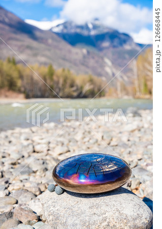 Steel tongue drum resting on a stone by the river in the Altai mountains. Concept of elemental harmony, raw sound energy, and nature as a sacred space for music and silence 126668445