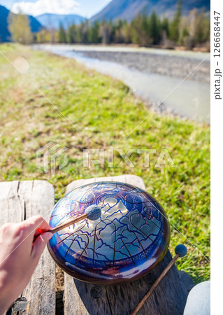 Point of view of person playing a steel tongue drum with mallets in front of mountains and river. Concept of immersive music, self-expression, and unity with natural rhythm 126668447