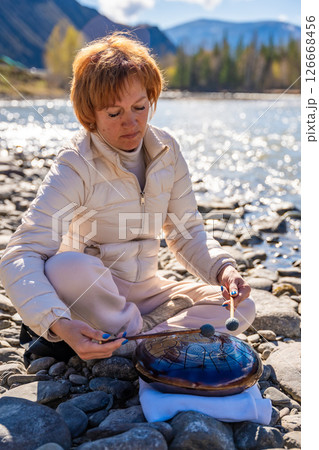 Woman playing a steel tongue drum near a mountain river in Altai, surrounded by wild nature. Concept of spiritual retreat, sound healing, and ancient connection to earth 126668456