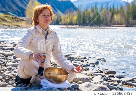 Woman performing a sound ritual with a Tibetan singing bowl by the Altai river. Concept of feminine energy, sacred vibration, and shamanic connection to the Earth Woman performing a sound ritual with a Tibetan singing bowl by the Altai river. Concept of feminine energy, sacred vibration, and shamanic connection to the Earth 126668458