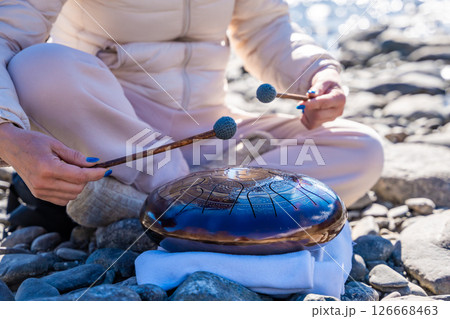 Close-up of hands playing a steel tongue drum by a mountain river in Altai. Concept of mindful music, meditative rhythm, and aesthetic harmony with wild nature. 126668463