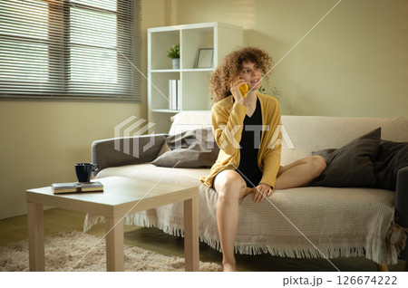 Curly haired woman in a mustard cardigan enjoys a cheerful phone call in living room 126674222