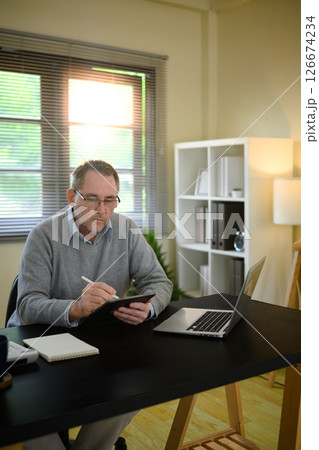 Focused older man writing on digital tablet with a stylus at his desk Focused older man writing on digital tablet with a stylus at his desk 126674234