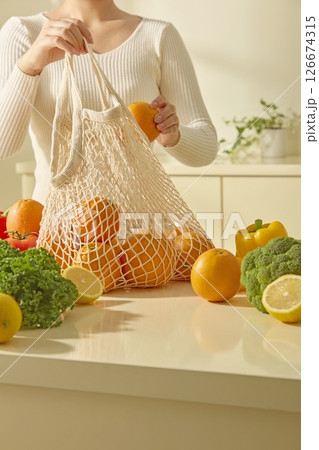 A woman places a reusable bag of fresh fruits and vegetables on a kitchen island, surrounded by colorful produce such as apples, celery, cauliflower, tomatoes and lemons. Kitchen background A woman places a reusable bag of fresh fruits and vegetables on a kitchen island, surrounded by colorful produce such as apples, celery, cauliflower, tomatoes and lemons. Kitchen background 126674315