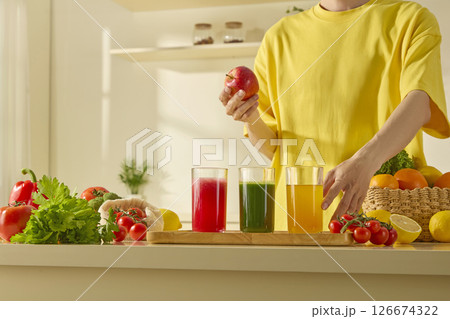 A person in a yellow T-shirt reaches for fresh vegetables and juices on a clean kitchen counter filled with healthy, colorful produce. Modern, sunny kitchen space A person in a yellow T-shirt reaches for fresh vegetables and juices on a clean kitchen counter filled with healthy, colorful produce. Modern, sunny kitchen space 126674322