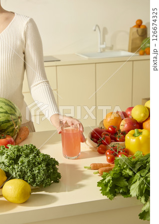 A woman is placing a glass of watermelon juice on a kitchen island filled with colorful vegetables: lemons, apples, tomatoes, celery, cauliflower... Minimalist, modern kitchen space 126674325