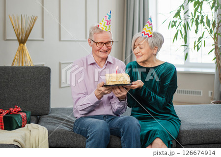 Make a wish. Family senior couple in party hat cap celebrating birthday anniversary together at home. Old woman blowing out burning candles on birthday cake. Old man wishes wife happy birthday Make a wish. Family senior couple in party hat cap celebrating birthday anniversary together at home. Old woman blowing out burning candles on birthday cake. Old man wishes wife happy birthday 126674914