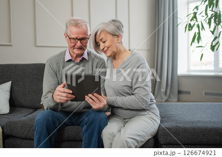 Senior old couple with tablet watching video online. Husband wife spending time together at home. Happy modern mature senior couple using tablet for family entertainment. Old people modern technology Senior old couple with tablet watching video online. Husband wife spending time together at home. Happy modern mature senior couple using tablet for family entertainment. Old people modern technology 126674921