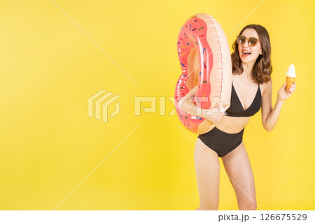 Cheerful Caucasian model in swimsuit hold soft serve cone and donut ring float on vibrant yellow studio backdrop with blank space ideal for lifestyle marketing, tropical travel, or dessert campaigns 126675529