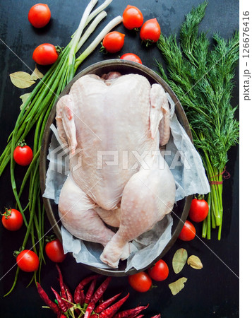 Whole raw chicken laying in the pan with parchment on the kitchen table preparing for baking, surrounded by fresh vegetables and herbs, top view. Whole raw chicken laying in the pan with parchment on the kitchen table preparing for baking, surrounded by fresh vegetables and herbs, top view. 126676416