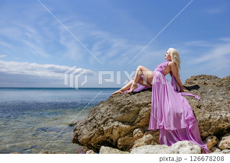 Woman beach dress: Blonde model poses on rocks by sea during daytime for photoshoot. Woman beach dress: Blonde model poses on rocks by sea during daytime for photoshoot. 126678208