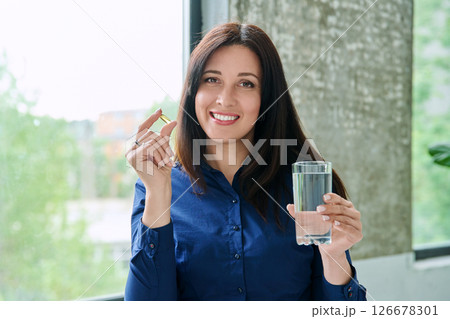 Beautiful woman taking omega 3 capsule fish oil pill, looking at camera with glass of water 126678301