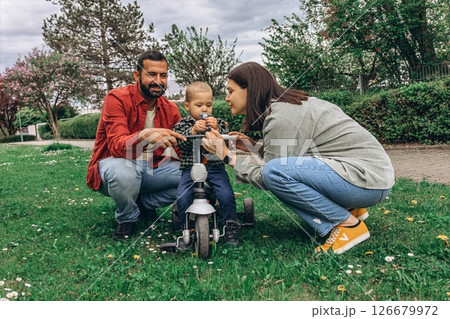 A mom and dad with their 2-year-old son in the park. The toddler is riding a small bike, enjoying family time outdoors filled with joy and learning 126679972