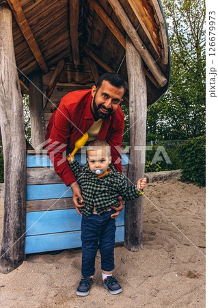 Father and young child playing together in a wooden playhouse at an outdoor playground. The child pours sand from a yellow scoop, surrounded by natural wood textures and greenery 126679973