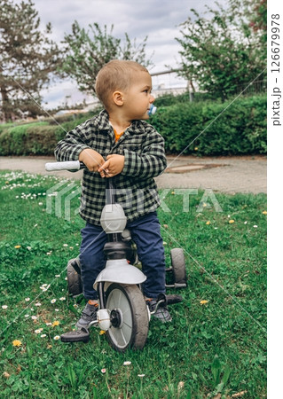 A 2-year-old boy riding a small bike in the park with a pacifier in his mouth. A sweet and candid moment of early childhood, outdoor activity, and toddler development in a natural setting 126679978