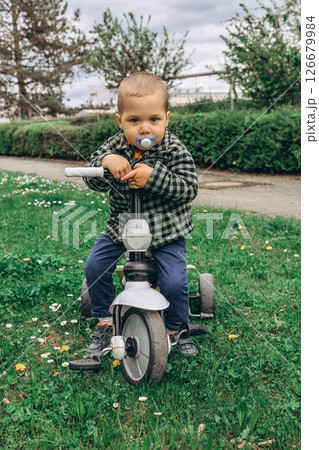 A 2-year-old boy riding a small bike in the park with a pacifier in his mouth. A sweet and candid moment of early childhood, outdoor activity, and toddler development in a natural setting 126679984