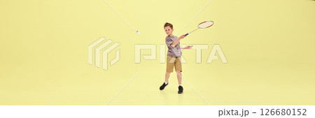 Boy lunges forward swinging racket at flying shuttlecock, fully immersed in game against pastel yellow background with copy space. Banner 126680152