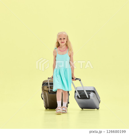 Girl in turquoise dress walks forward pulling two rolling suitcases, looking straight with confident smile against clean yellow studio background. 126680199