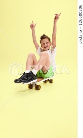 Child sitting on skateboard with raised arms, smiling widely and showing excitement, dressed in green shorts and white tee against yellow background 126680206