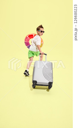 Boy walking forward and looking back while pulling suitcase, holding inflatable swim ring, ready for beach adventure against yellow background 126680223