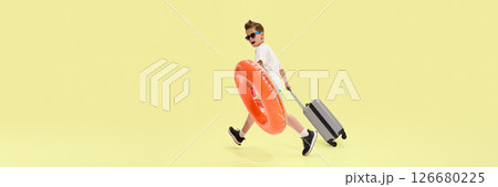 Playful boy in sunglasses walking with suitcase and swim ring, preparing for summer journey with happy and carefree attitude against yellow background 126680225