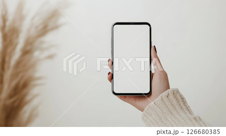Woman holding a smartphone featuring a blank white screen, positioned against a clean white wall and pampas grass, creating an ideal setting for mockup design and digital presentations 126680385