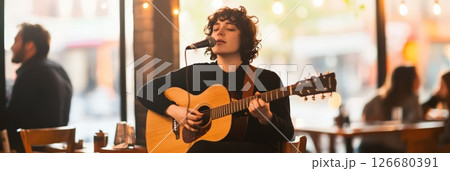 Young woman with curly hair singing and playing an acoustic guitar during a live music performance in a cozy restaurant, creating a warm and intimate atmosphere for the patrons enjoying their meals 126680391