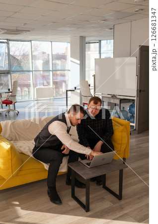 Two Caucasian men discussing work on a laptop while sitting on a sofa. Modern office. Two Caucasian men discussing work on a laptop while sitting on a sofa. Modern office. 126681347